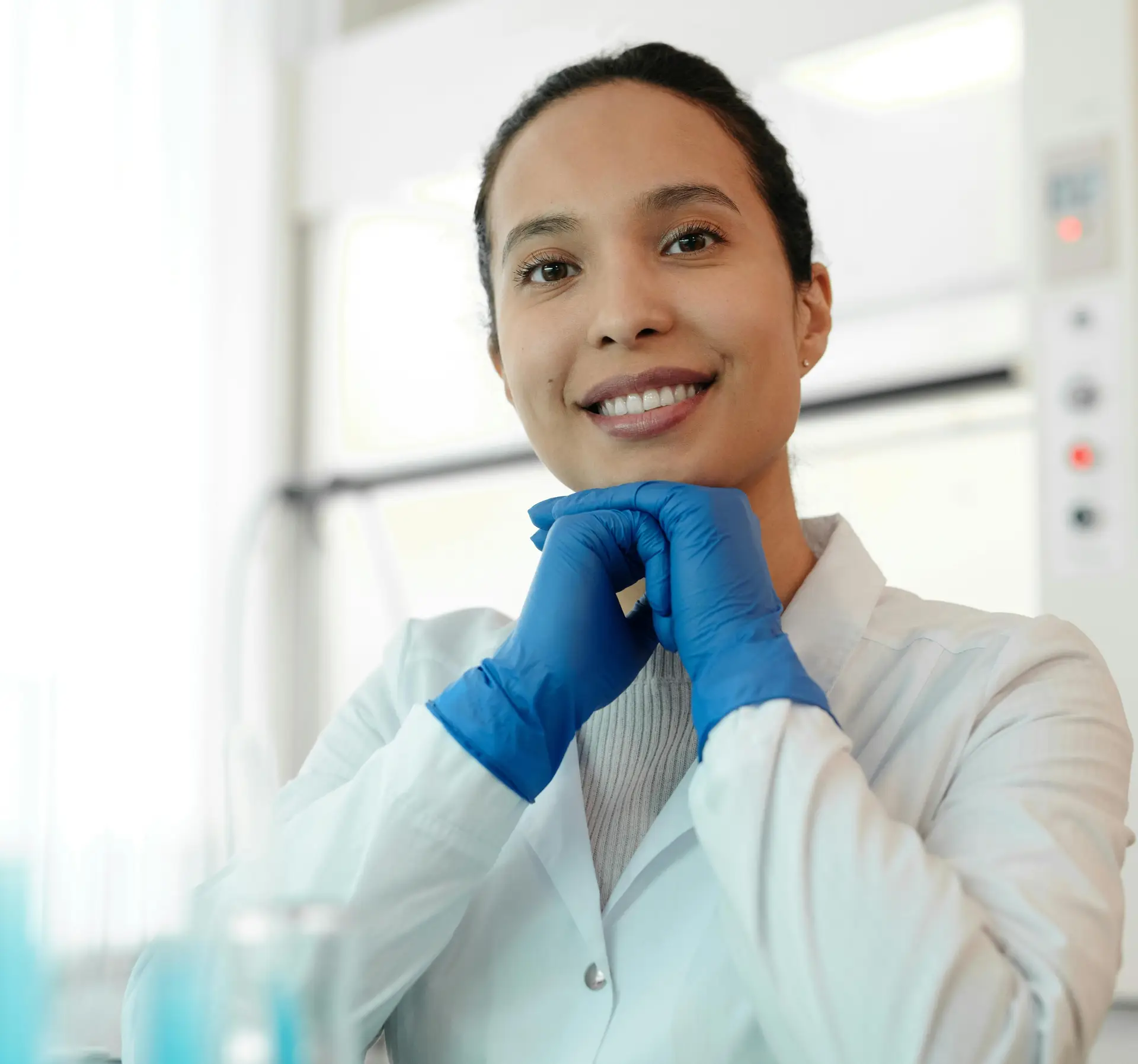 A smiling female scientist in a white lab coat, sitting in a laboratory with test tubes.